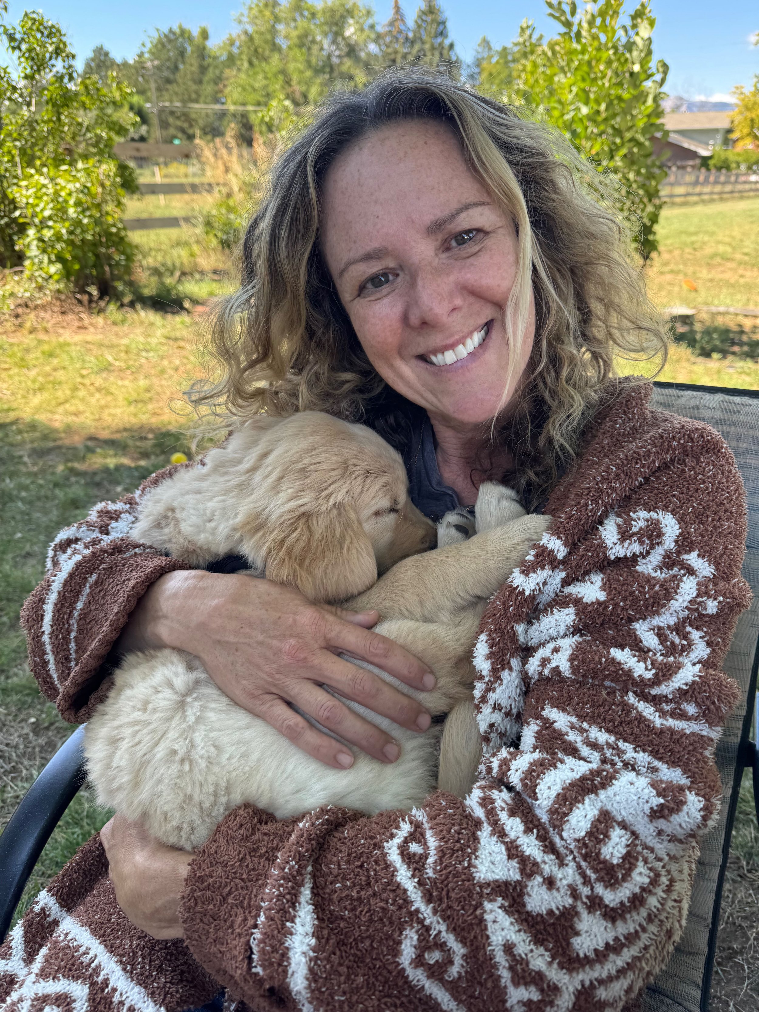 A woman with curly blonde hair and freckles holding a golden retriever puppy outdoors on a sunny day, with trees and a fence in the background.