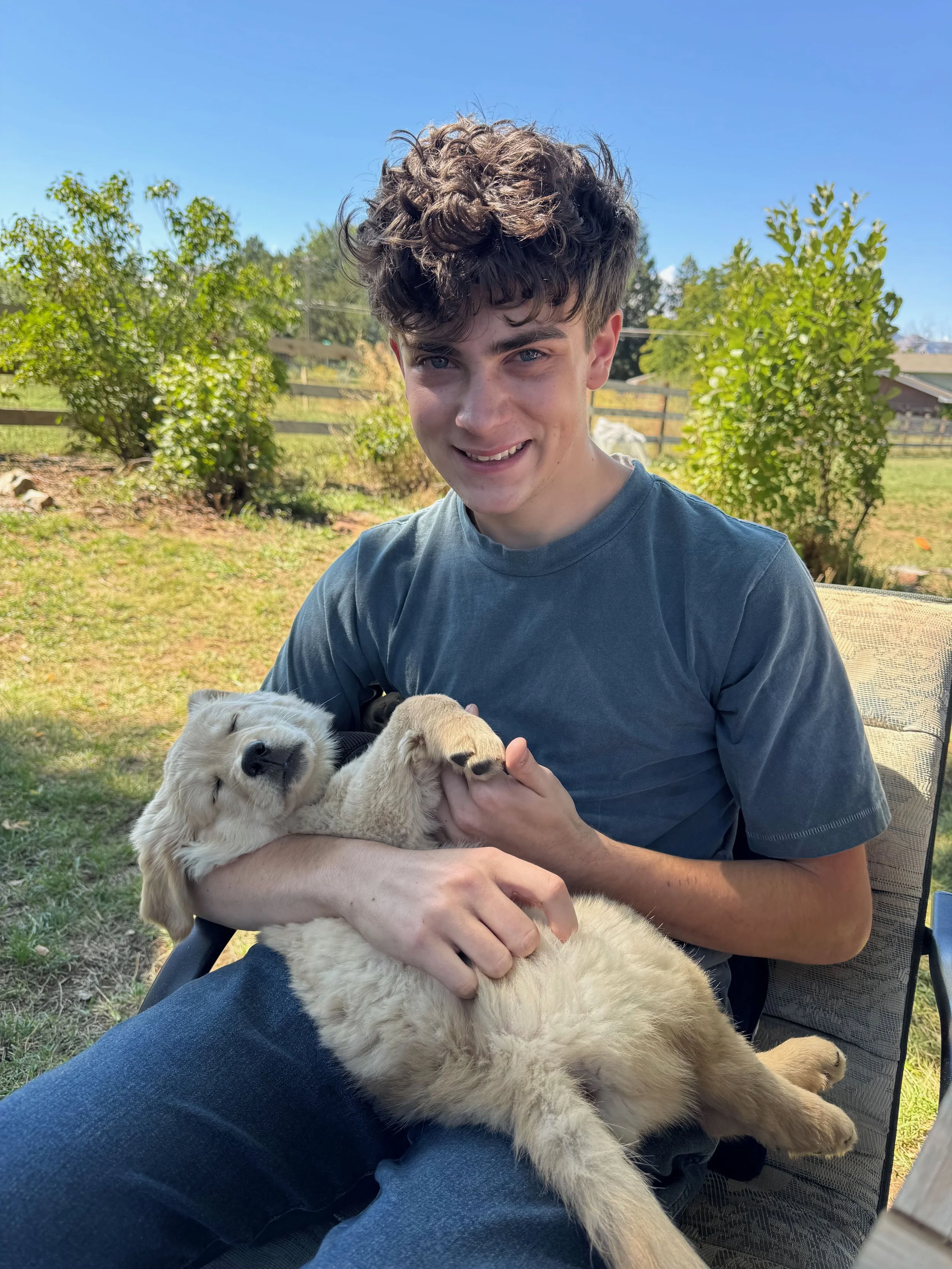 A young man with curly brown hair and blue eyes smiling while holding a fluffy light-colored puppy in his arms outside in a backyard with green bushes and a blue sky.
