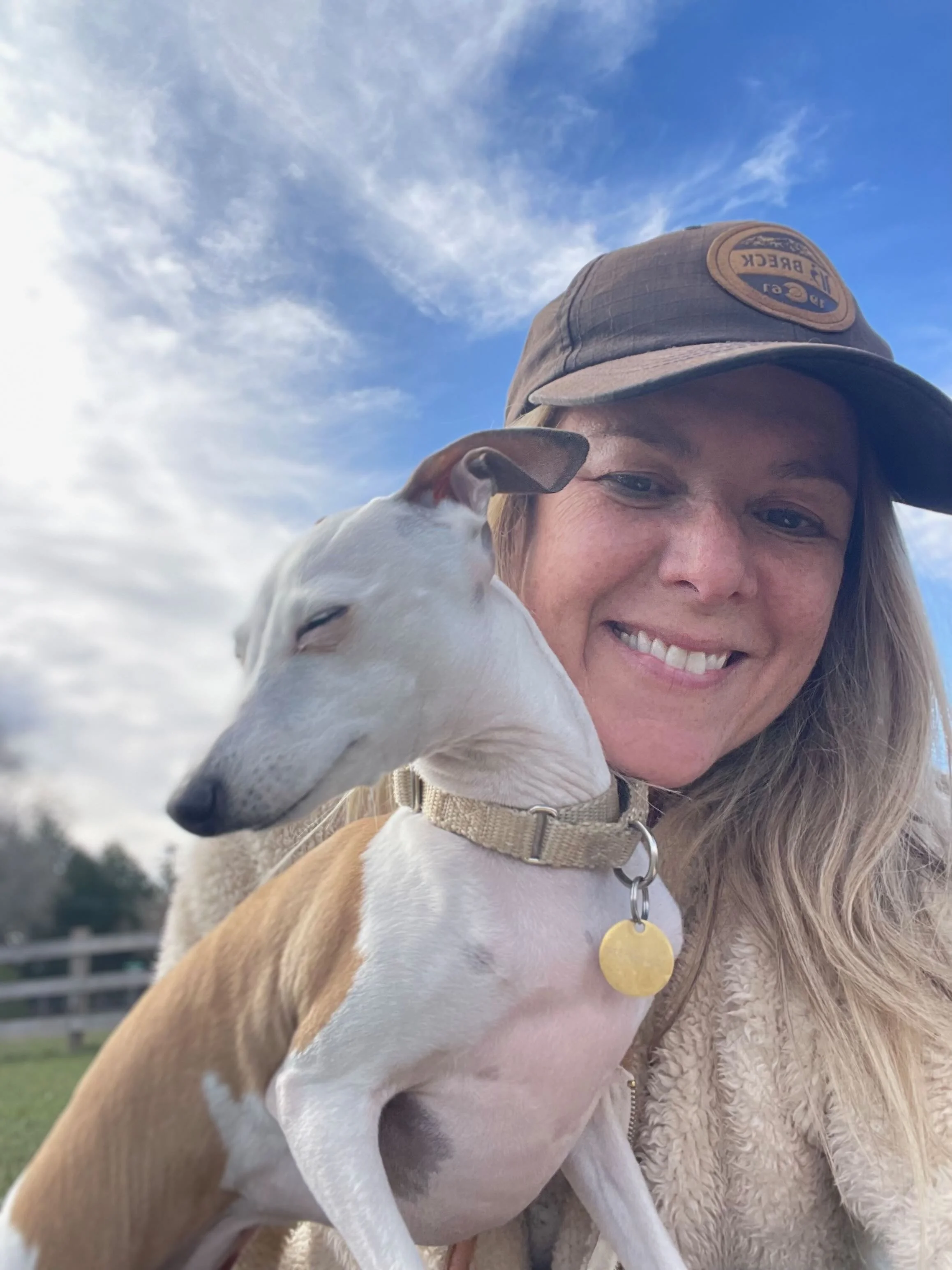 A woman smiling outdoors with a blue sky and clouds in the background, holding a white and brown dog with a tan collar and round gold tag.