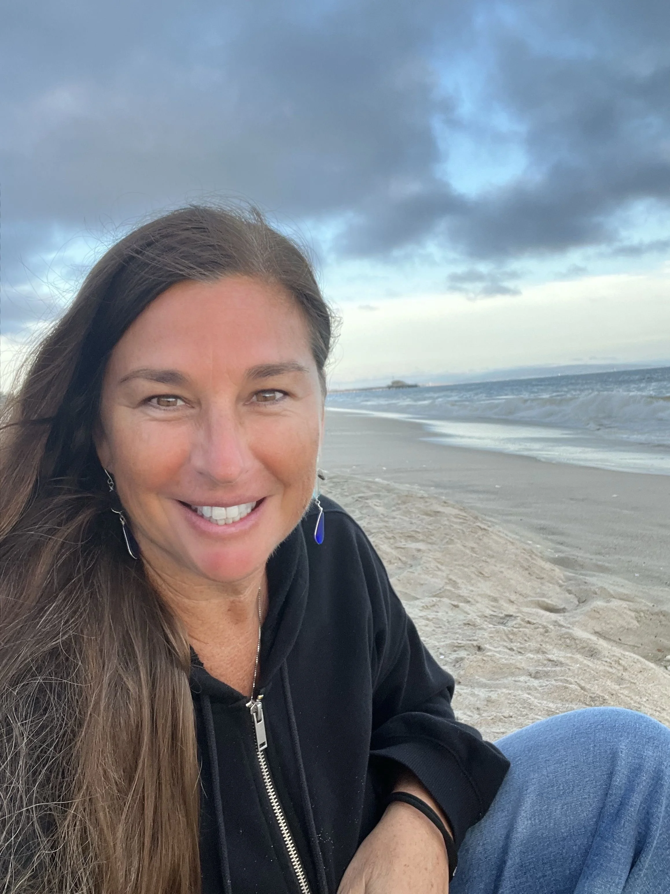 A woman with long brown hair smiling on a beach with ocean waves and cloudy sky in the background.