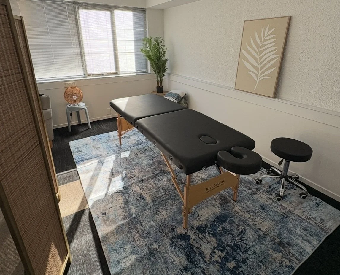 A massage therapy room with a massage table, a window with blinds, a decorative plant, a framed leaf print on the wall, a small stool, and a patterned rug.