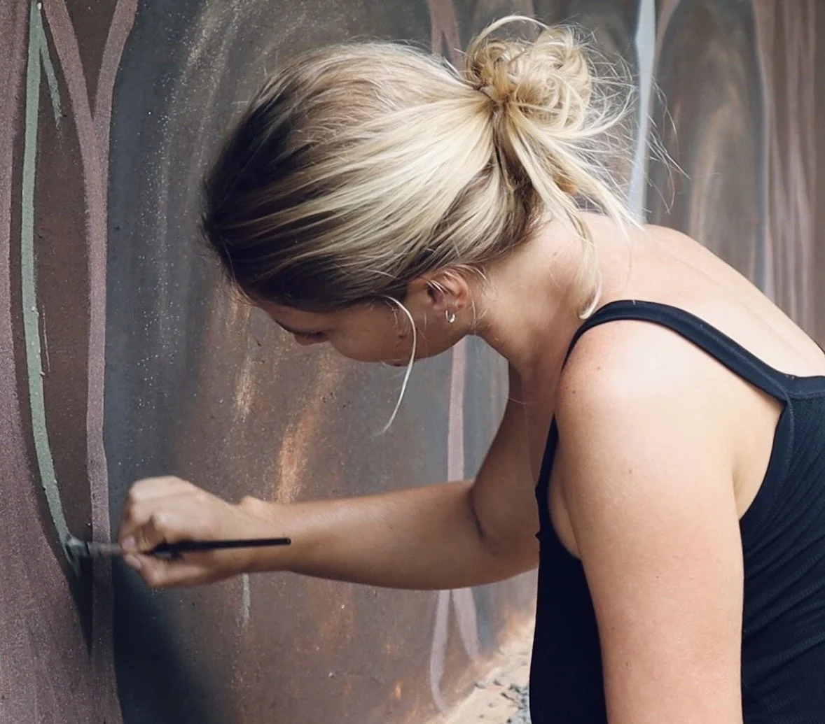 A woman with blonde hair in a bun, wearing a black tank top, is painting a mural on a wall with a small brush.