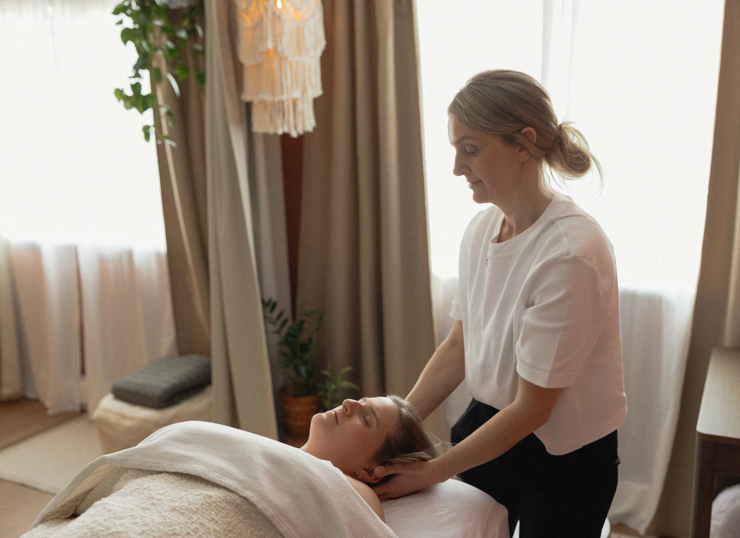A woman receives a massage from a massage therapist in a cozy, softly lit room decorated with plants, curtains, and a hanging textile art piece.
