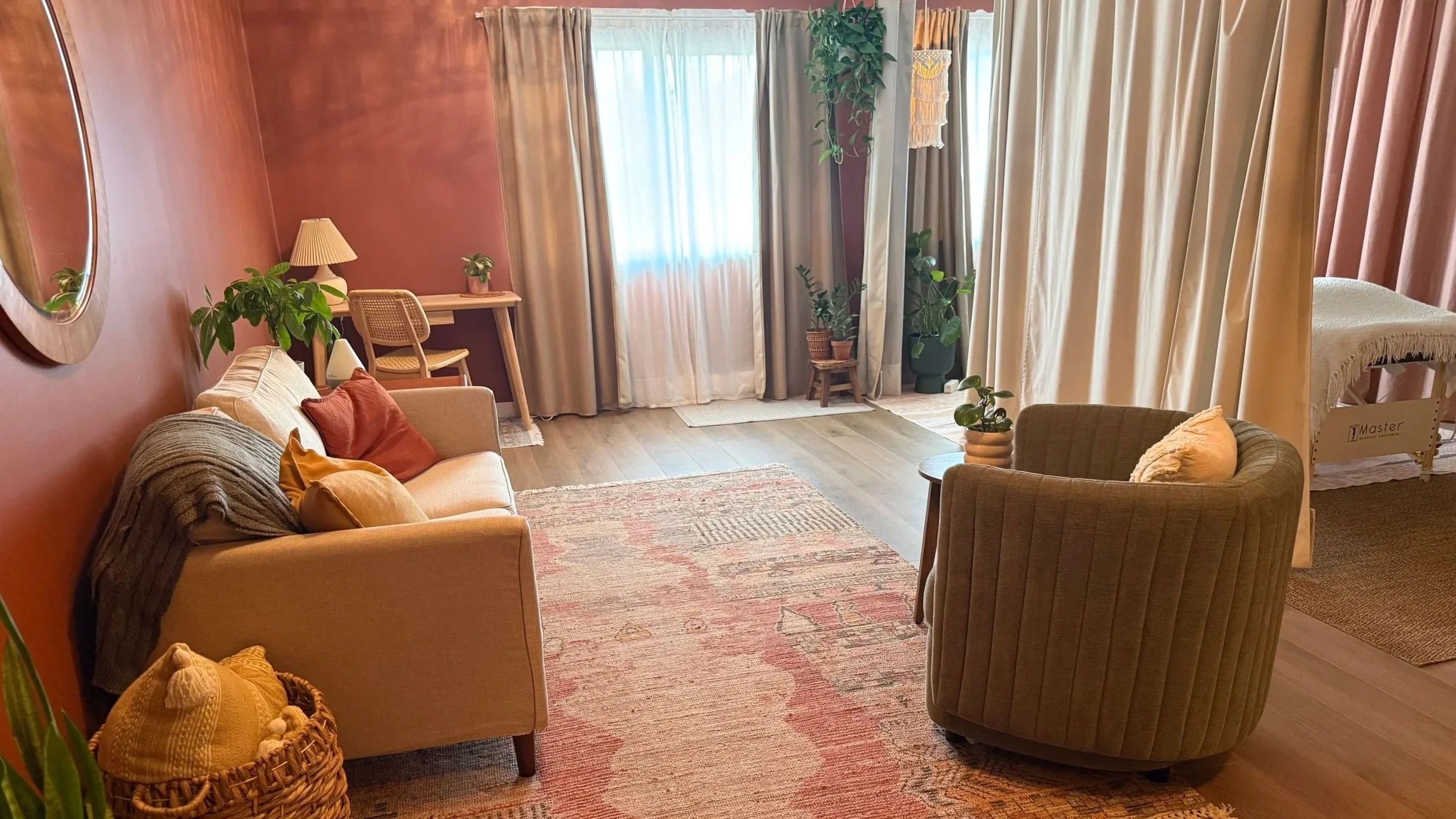Living room with pink walls, beige sofa with pillows, gray armchair, wooden side table, large window with curtains, potted plants, and a striped rug.