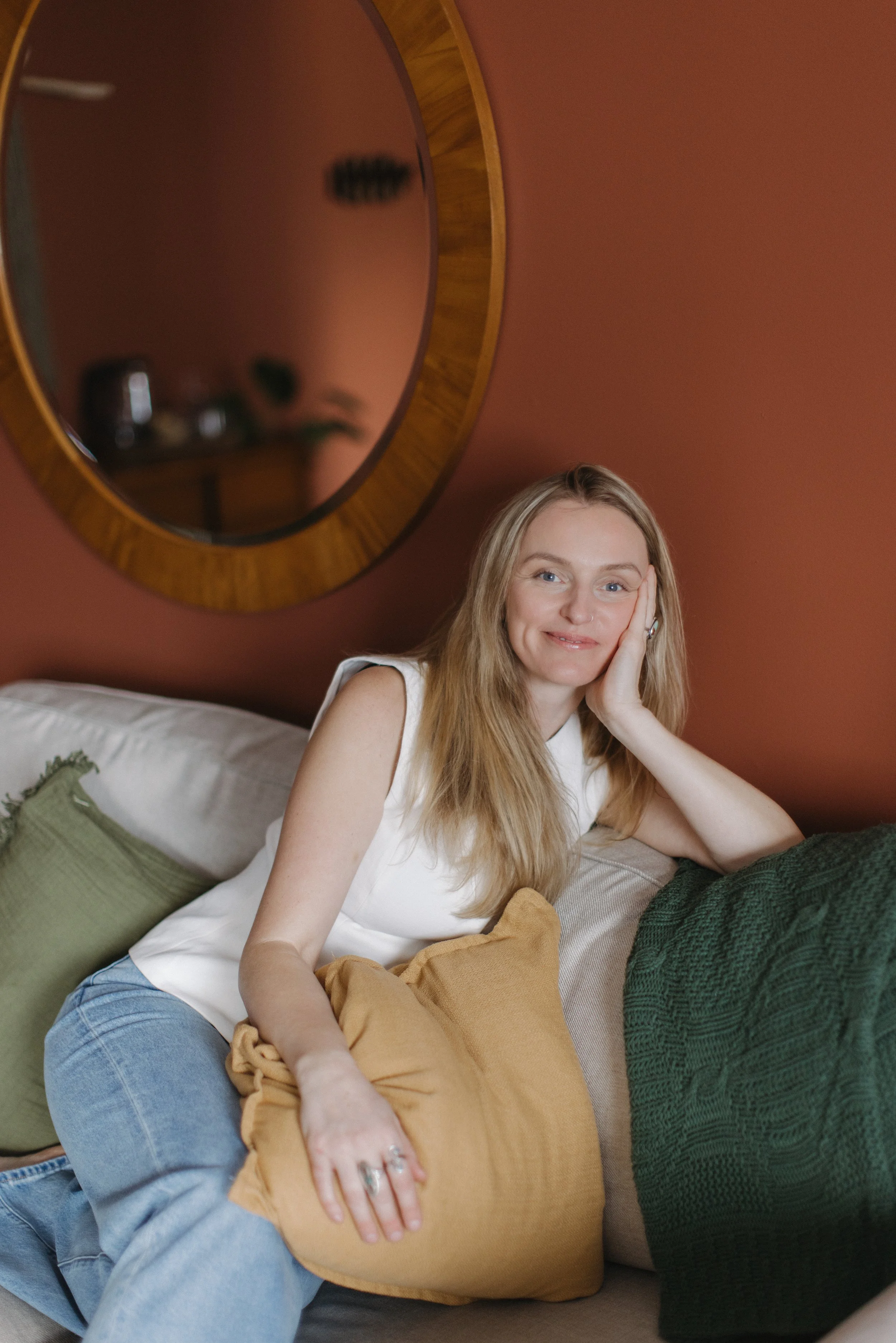 A young woman with long blonde hair, wearing a white sleeveless top and light blue jeans, sitting on a couch with a green pillow and a beige blanket. She has a gentle smile, resting her face on her hand, in a room with warm, earthy wall color and a round wooden mirror hanging on the wall behind her. The mirror reflects some furniture and objects.