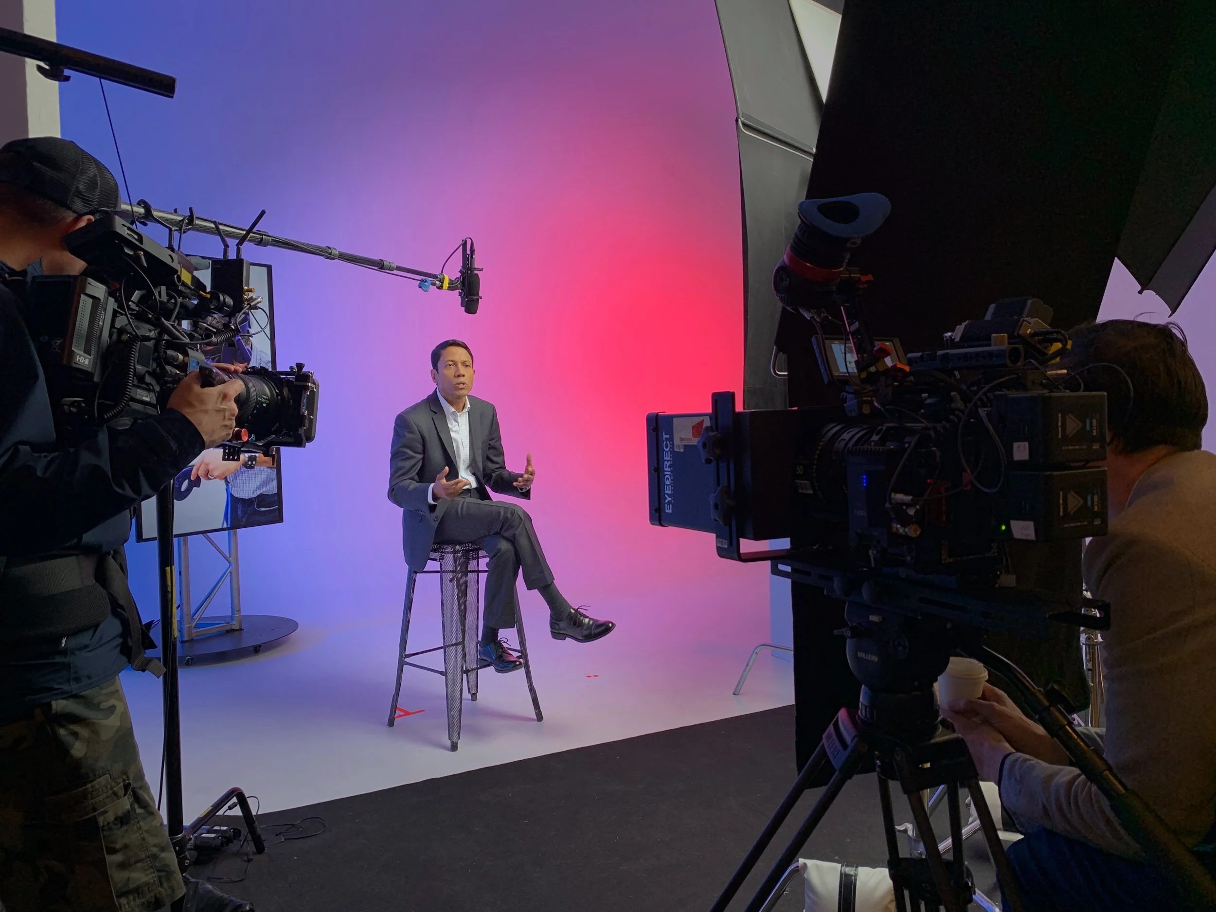 Anoop Chaudhuri in a suit sitting on a tall stool speaking during a recording session with camera crew and professional equipment, colourful background with gradients of purple and pink.