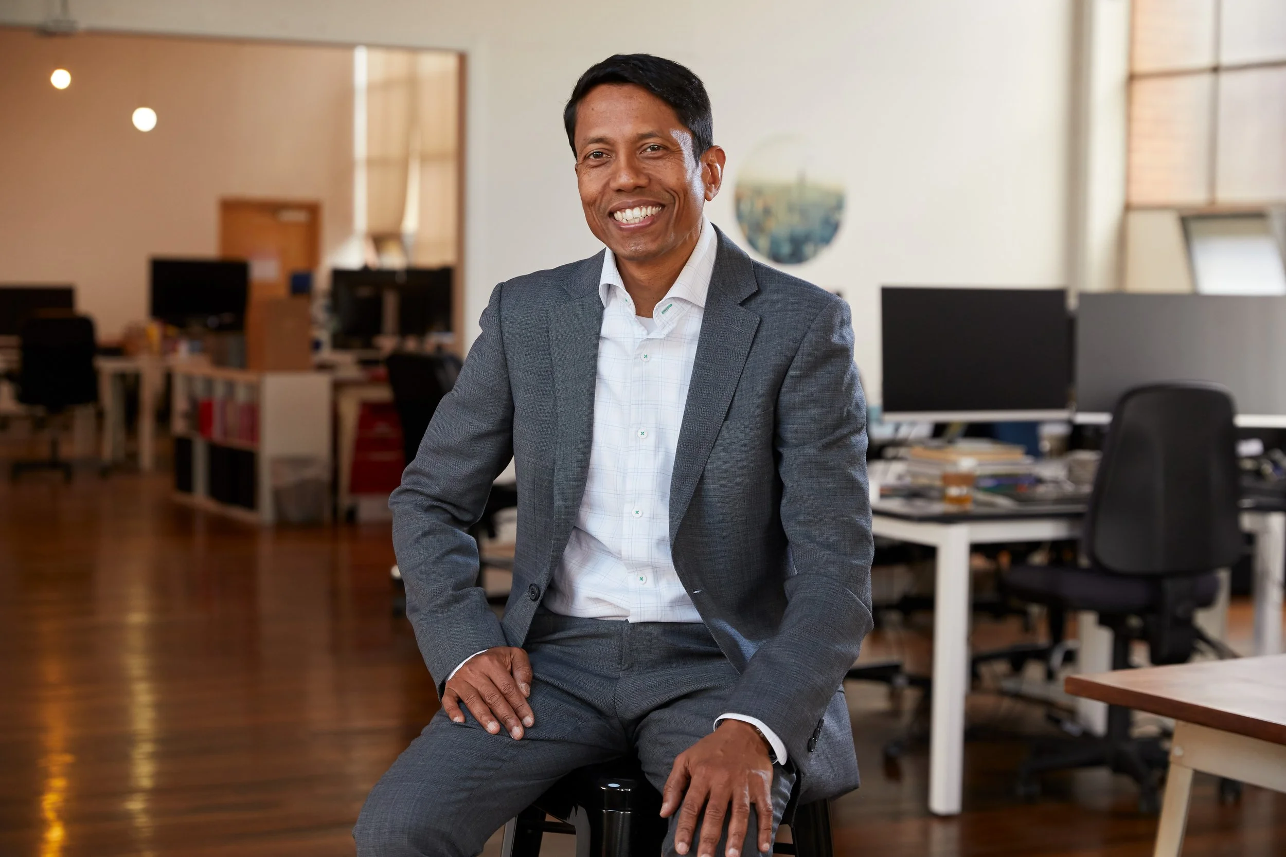 Anoop Chaudhuri in a grey suit and white shirt sitting in an office environment with computer desks and chairs in the background.