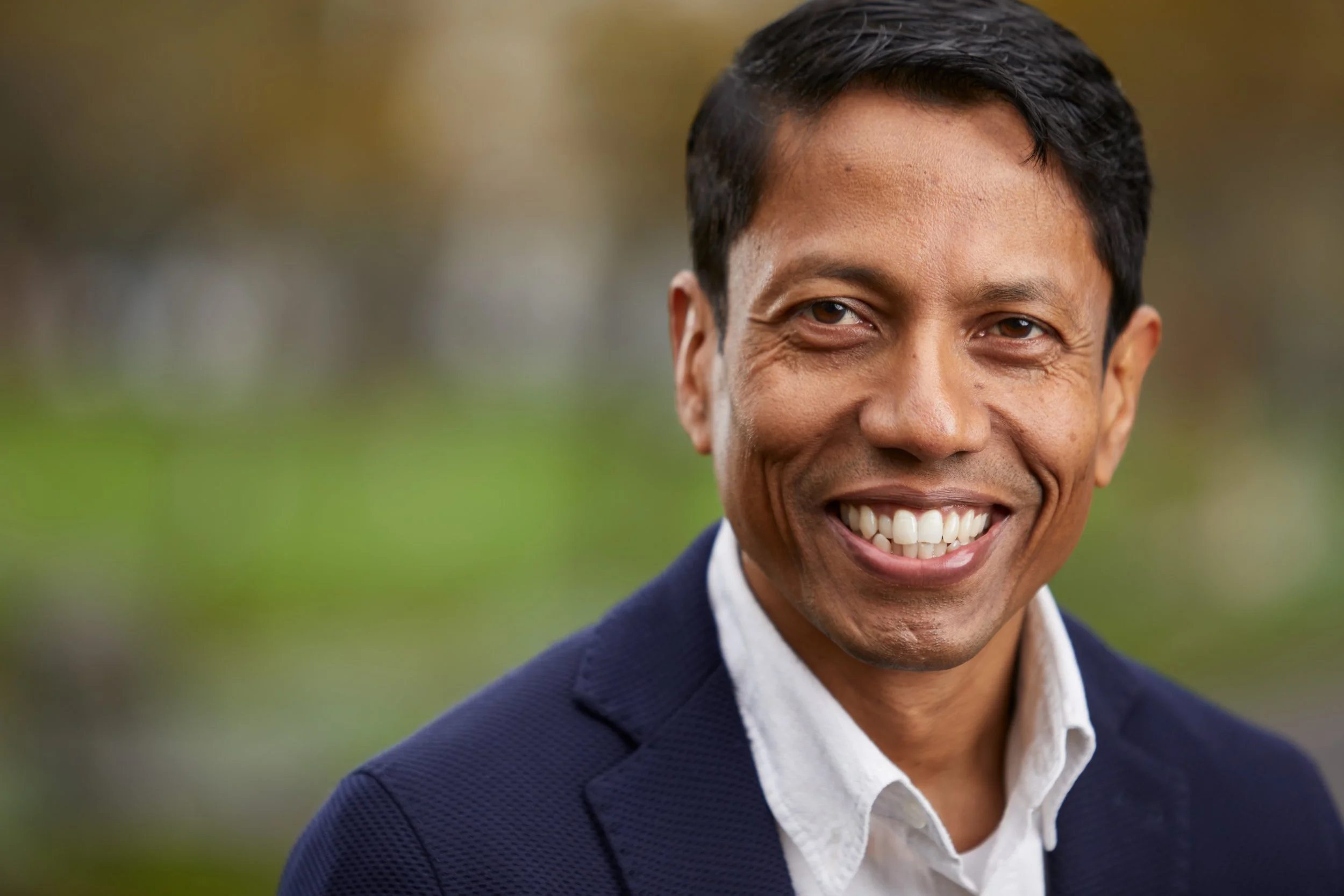 Close-up of Anoop Chaudhuri smiling. He has short black hair, is wearing a white shirt and a navy blue blazer, standing outdoors with a blurred background.