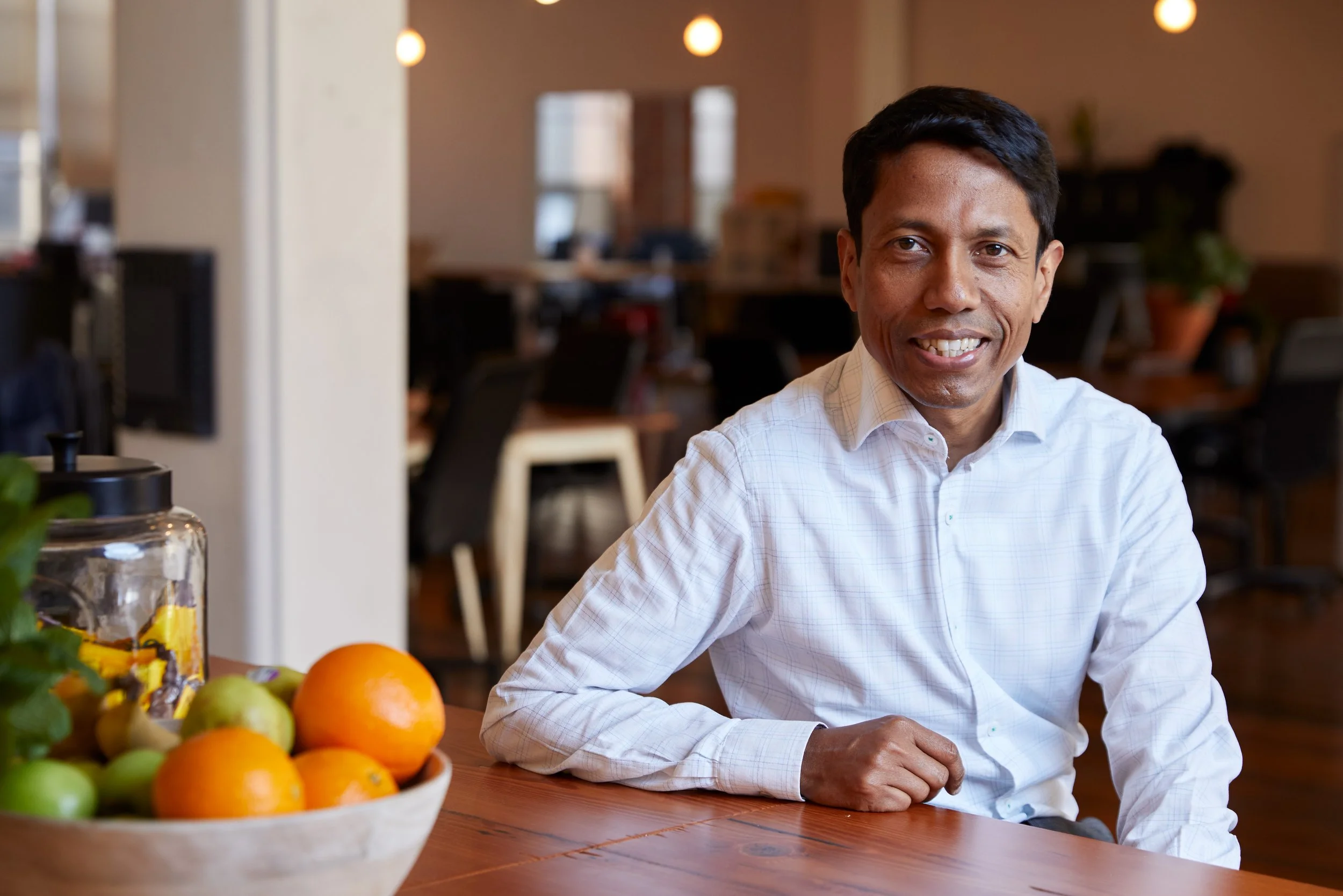 Anoop Chaudhuri in white shirt sitting at wooden table in modern office space with fruit bowl in foreground.