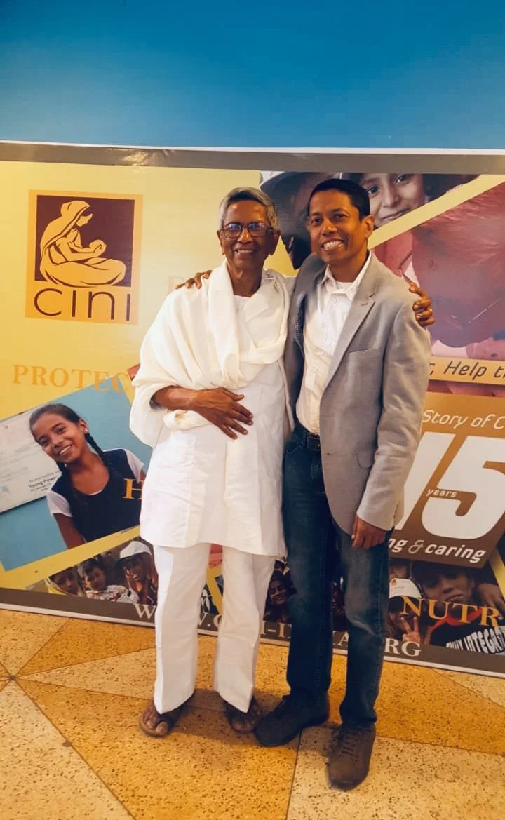 Two people, Anoop Chaudhuri man in a grey blazer and his father in traditional white clothing, smiling and standing close together in front of a colourful banner with the organisation logo 'cini' and images of children.