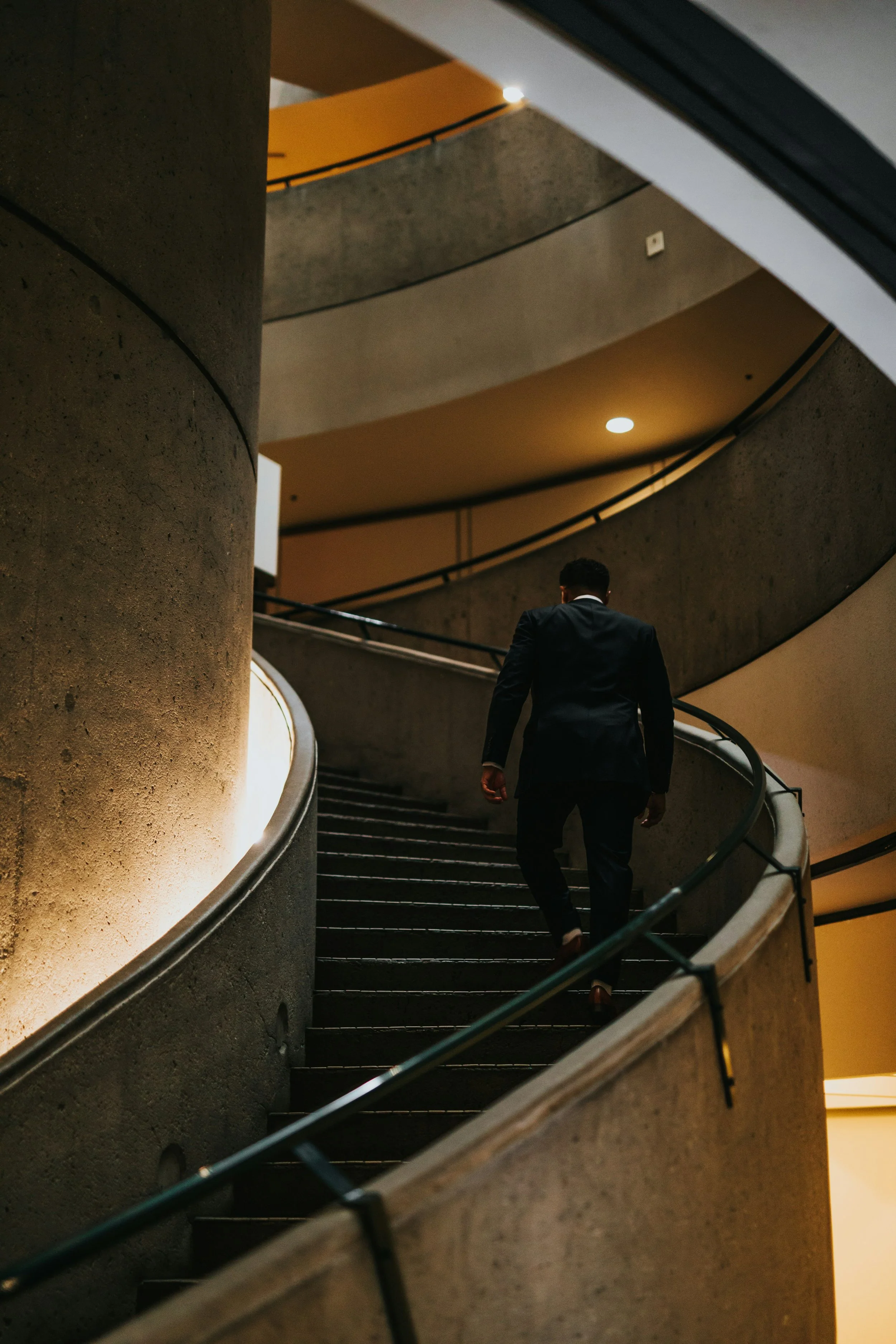 A man in a dark suit walking up a curving staircase inside a building with warm lighting.