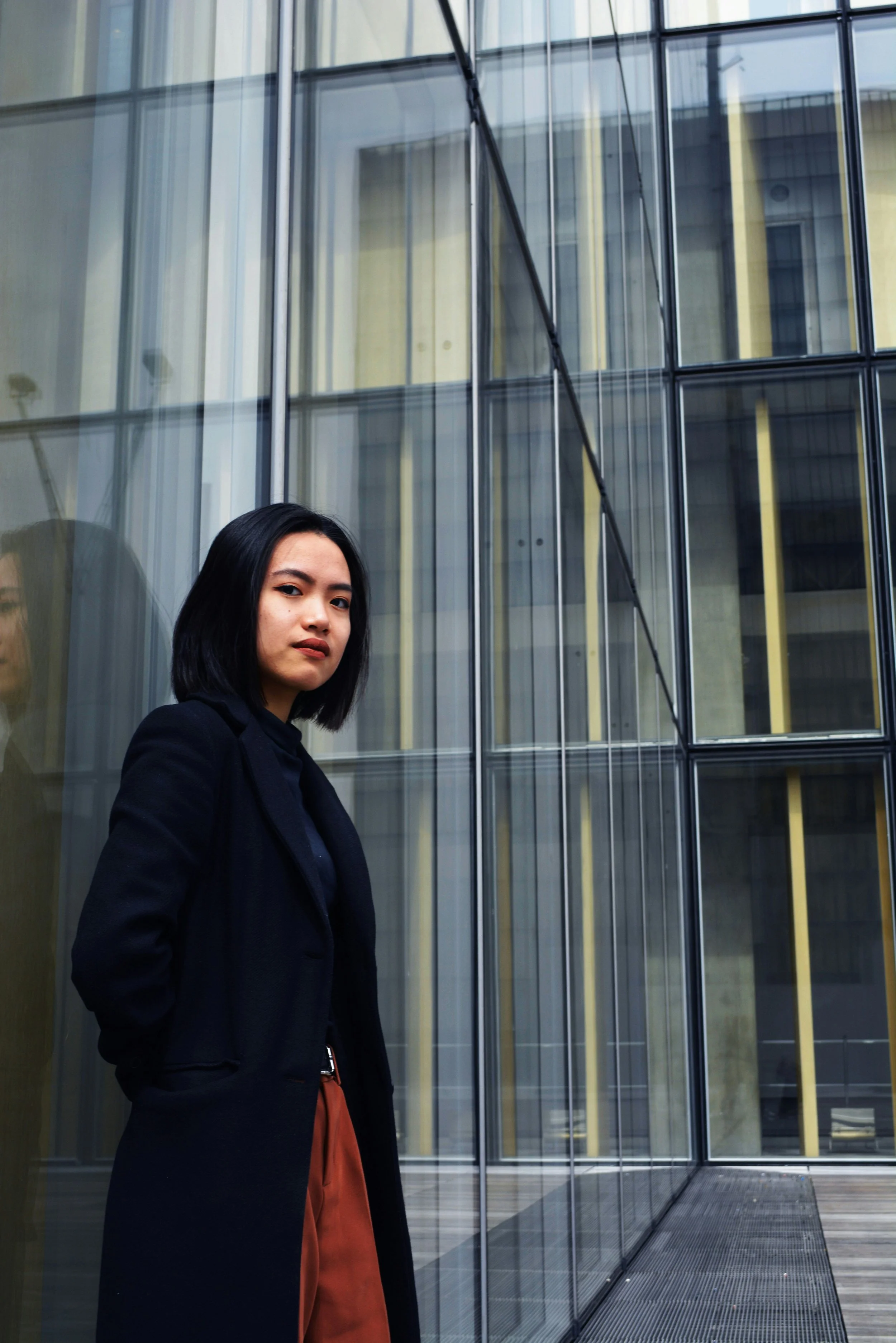 A woman standing outside near a modern glass building, with her arms crossed and wearing a black coat and reddish-brown pants.
