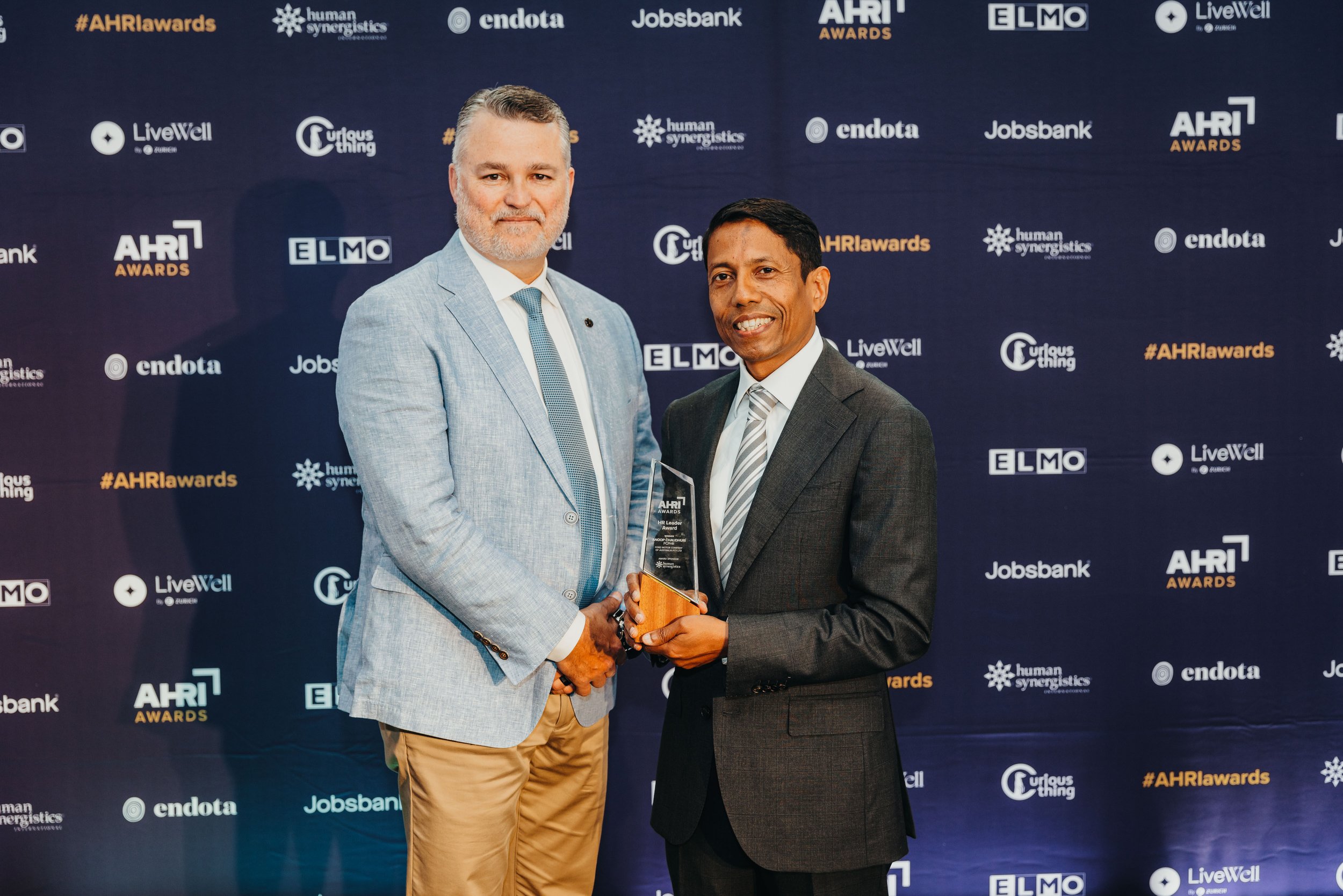 Two men in suits standing together at an awards ceremony, one is handing Anoop Chaudhuri an award. The background has logos and text related to HR awards and sponsors.