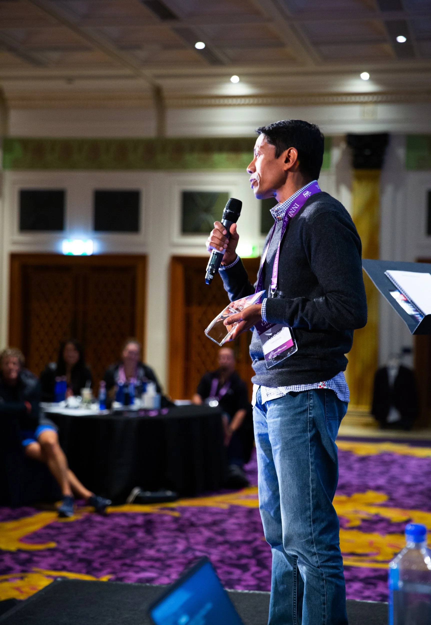 Anoop Chaudhuri in a black sweater and blue jeans speaks into a microphone during a conference. The background features an audience sitting at tables, in a decorated conference room with purple and yellow carpet and white walls.