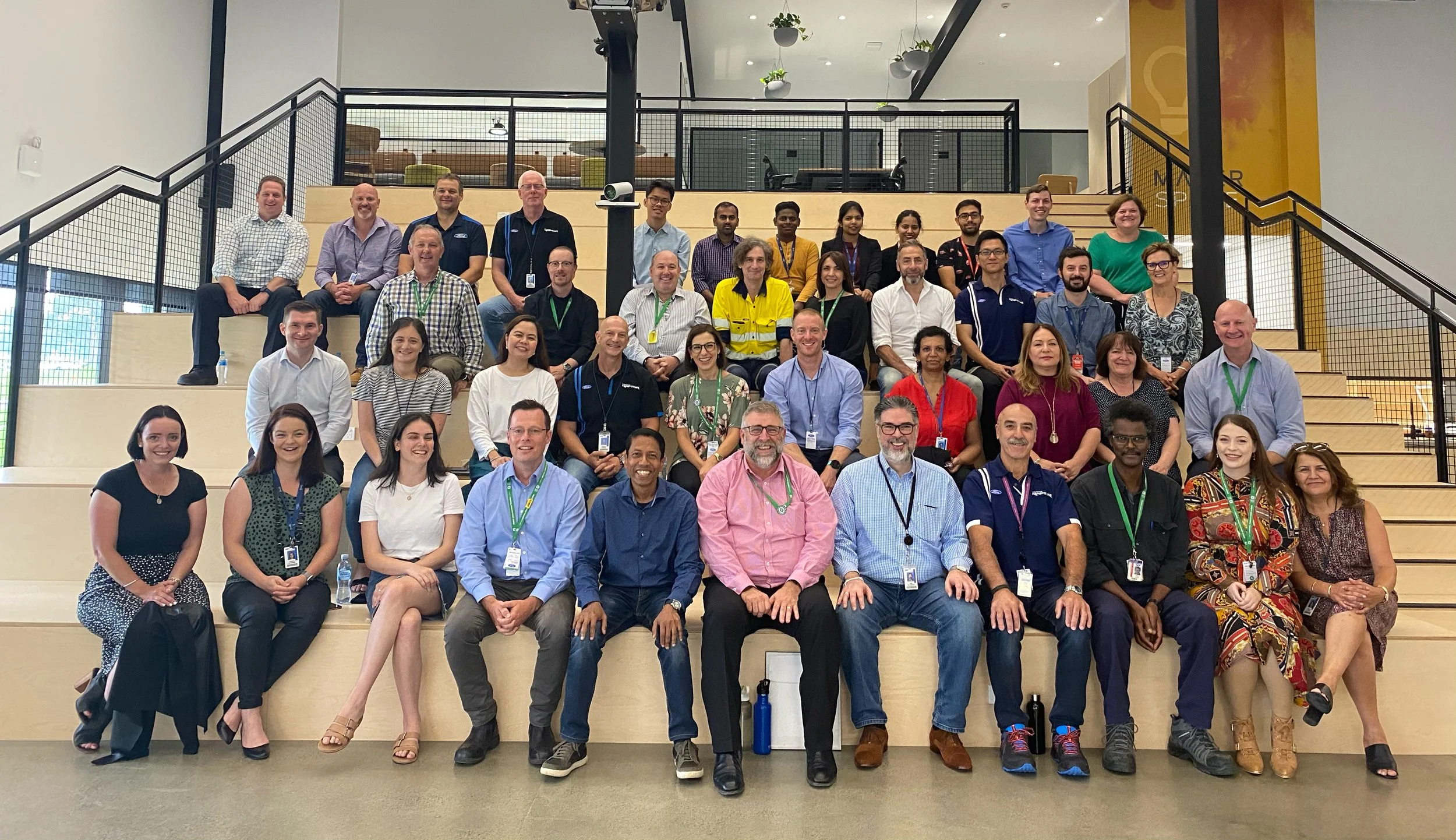 Group photo of diverse men and women sitting and standing on a wide staircase indoors, with some wearing lanyards and ID badges, in a modern building with plants hanging from the ceiling.