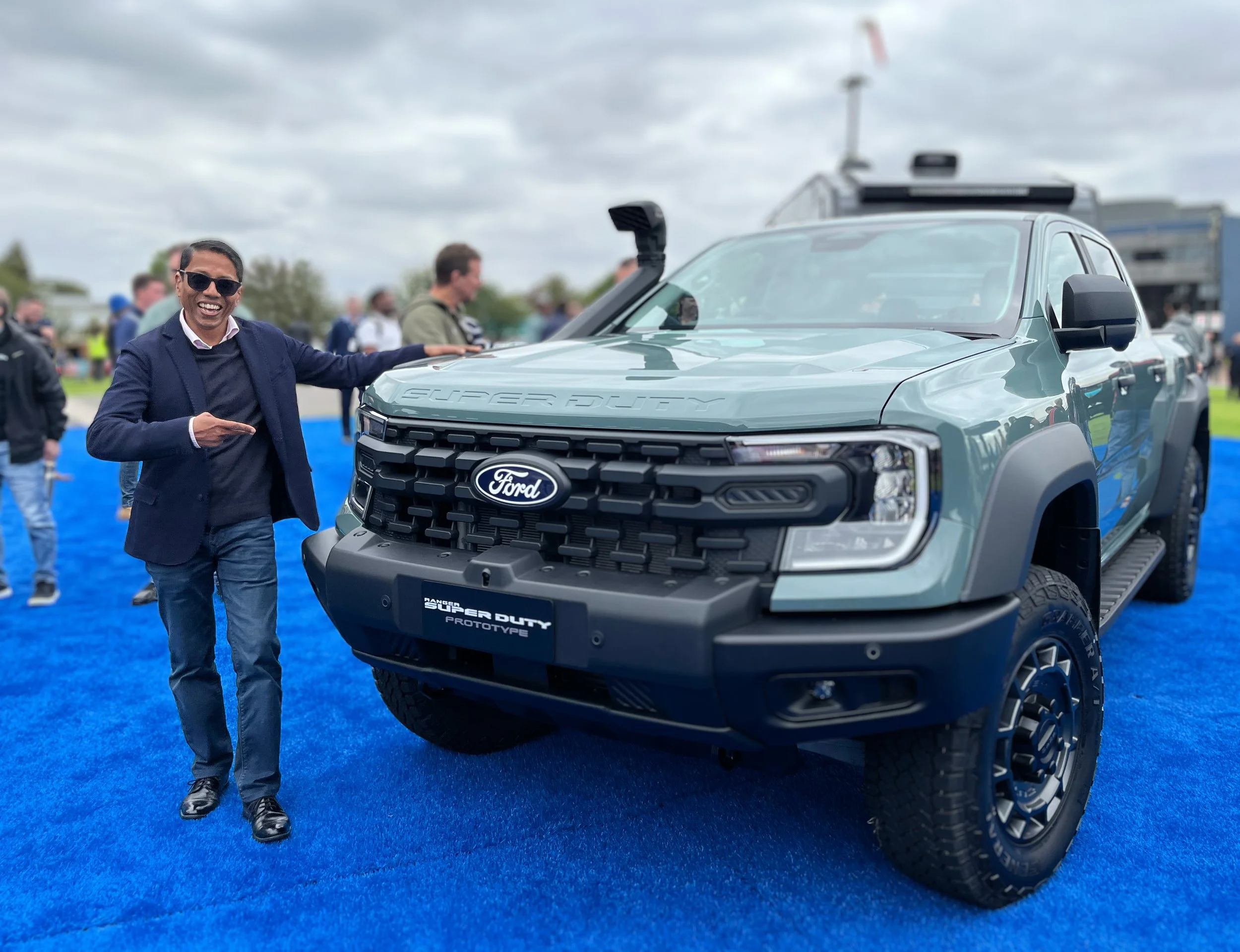 Anoop Chaudhuri wearing sunglasses and a suit jacket standing next to a blue Ford Super Duty prototype ute at an outdoor event with a cloudy sky and other people in the background.