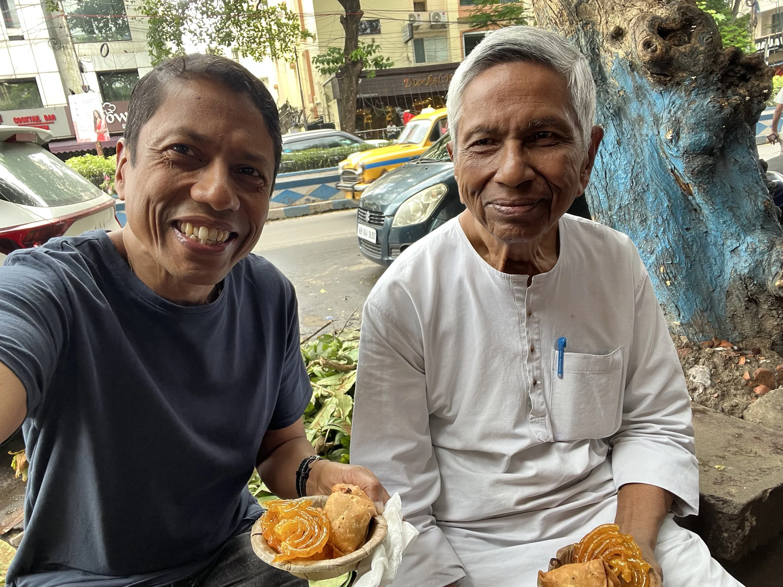 Two men sitting outdoors on a street, smiling. Anoop Chaudhuri is holding a basket with baked goods, including pastries and sweets. Behind them are cars, trees, and buildings.