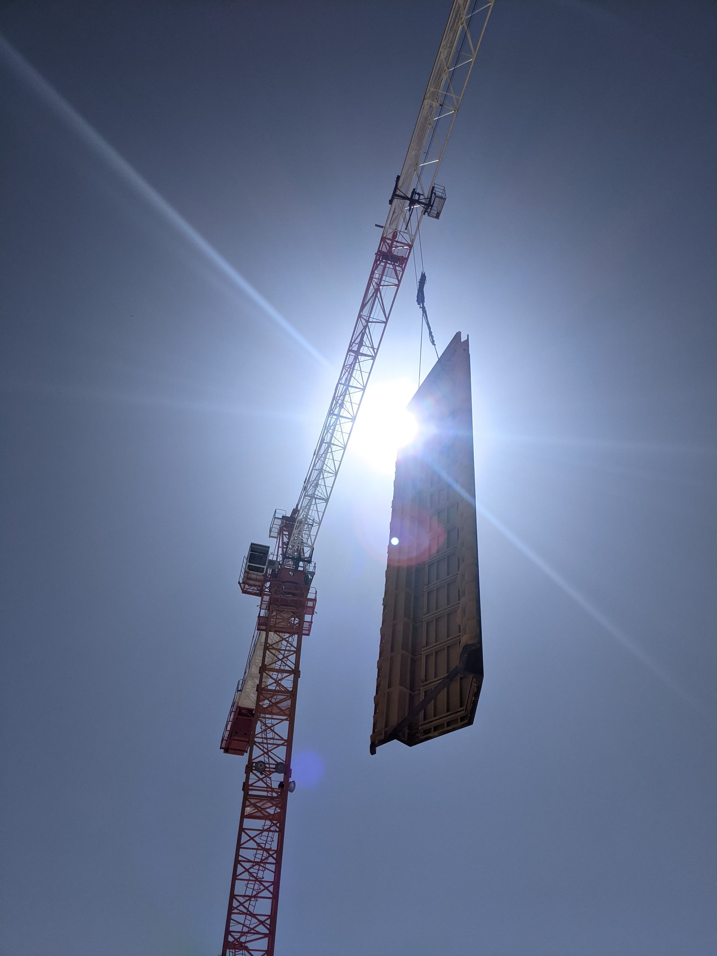 A construction crane lifting a building or large structure against a clear blue sky with the sun shining brightly behind the structure.