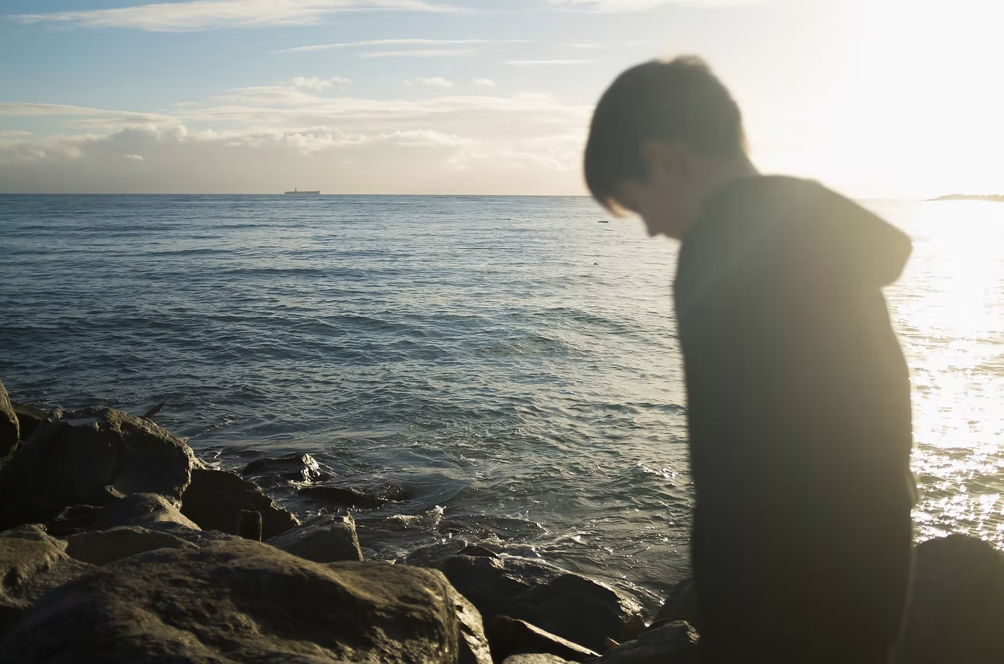 A person standing on rocks by the ocean during sunset, with the sun behind them and a ship visible on the horizon.