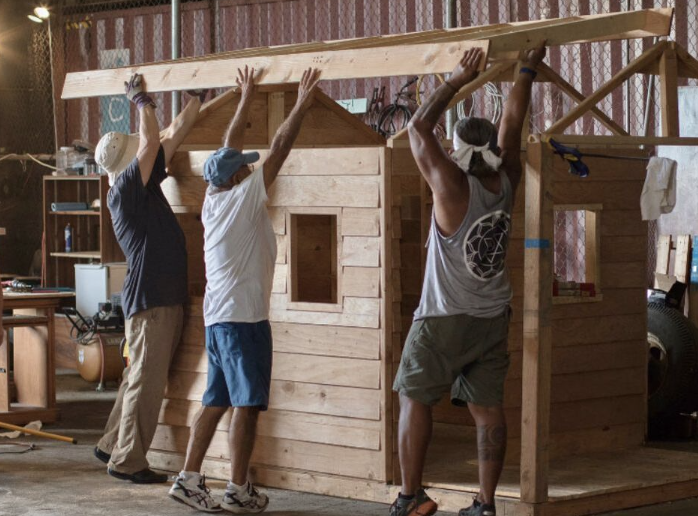 Three men working together to build a wooden shed or small house in a workshop or garage.