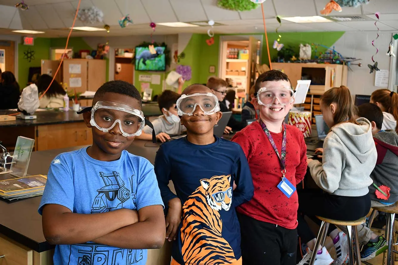 Three children wearing safety goggles in a classroom with other students working on laptops and books, decorated with hanging paper ornaments.