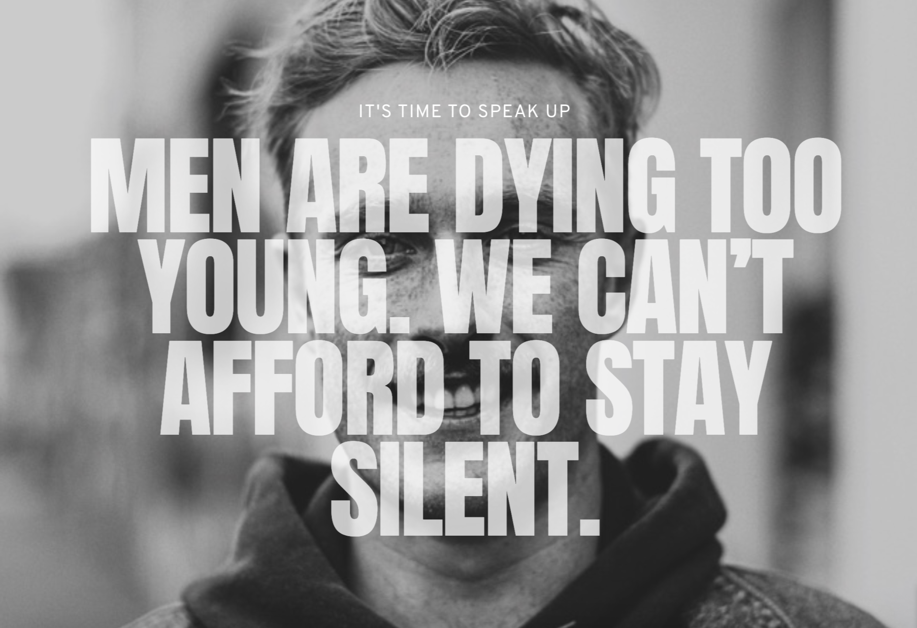 Black-and-white photo of a young man with freckles and short hair, smiling widely with his mouth open, showing his teeth, and wearing a dark hoodie. Overlaid large text reads: "Men are dying too young. We can't afford to stay silent."
