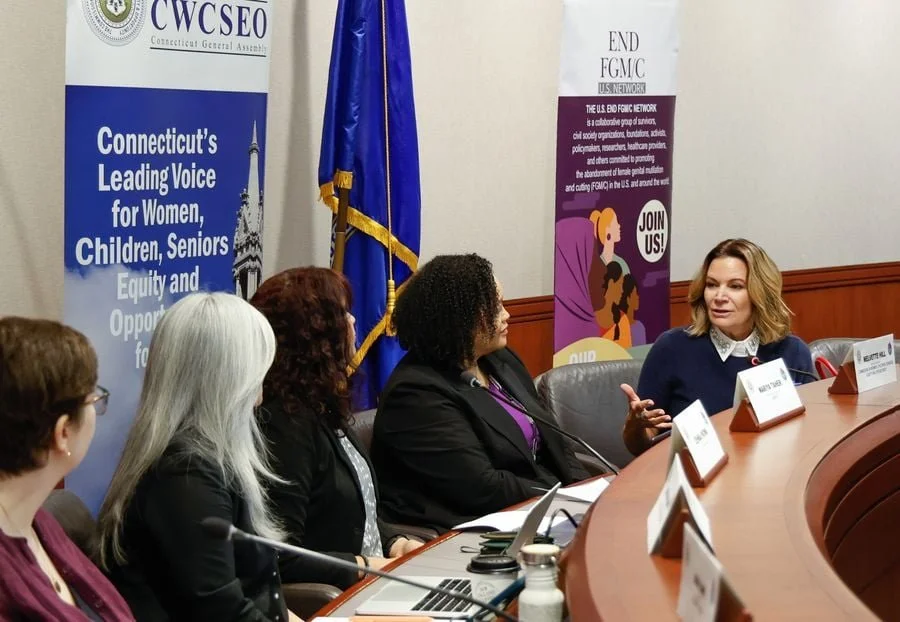 Women seated at a round conference table engaged in a discussion, with banners behind them promoting women's rights and advocacy at a formal event.