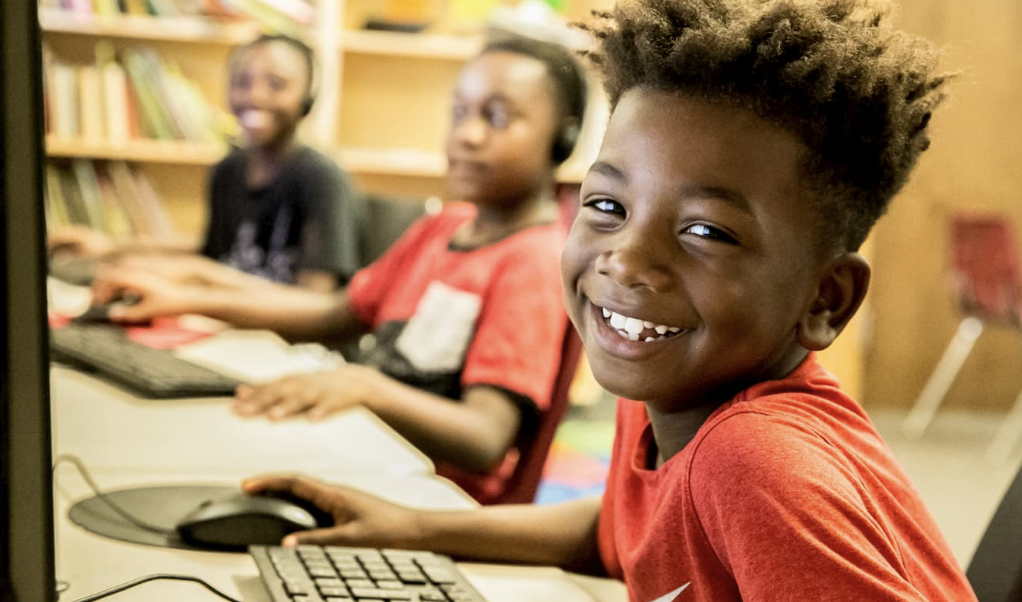 Three children using computers in a classroom, smiling and looking at the camera.