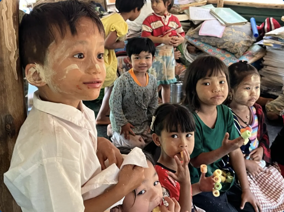 Group of children sitting on the floor inside a room, some with painted faces, holding colorful rings, with books and papers in the background.