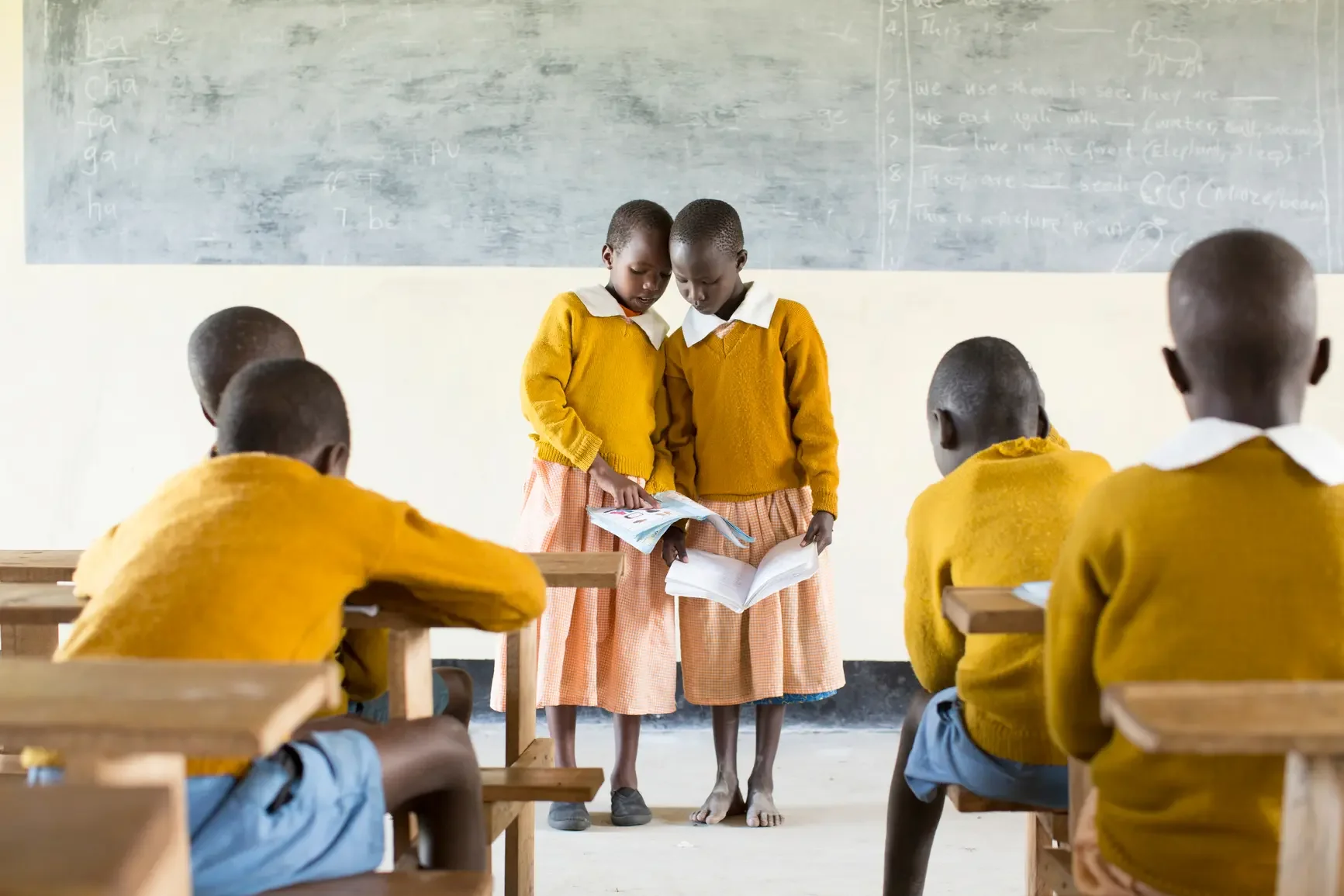 Two young girls in yellow sweaters and pink skirts standing in front of a classroom with boys in yellow sweaters sitting at wooden desks, looking at their books.
