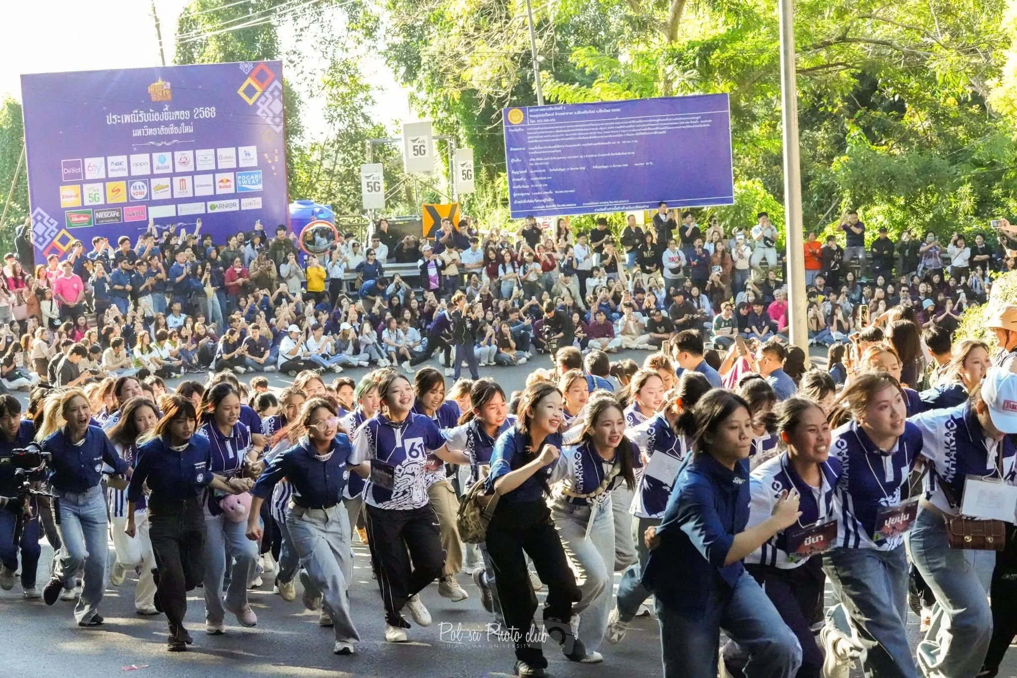 Large crowd of people, mostly young women, participating in a marathon or parade on a sunny day, with trees and large advertising boards in the background.