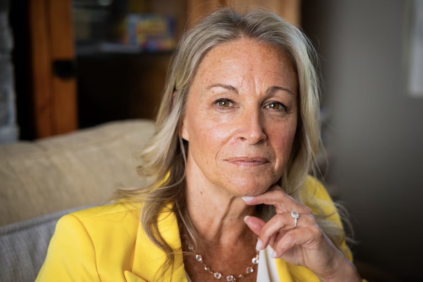 Close-up of a middle-aged woman with blonde hair wearing a yellow blazer, sitting on a beige couch with a thoughtful expression, hand resting on chin, in a cozy room with a bookshelf in the background.