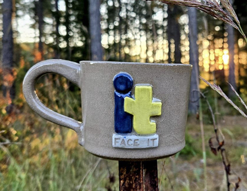 A ceramic mug with a colorful logo that says 'It' with the phrase 'Face It' underneath, set outdoors with a forest and sunset in the background.
