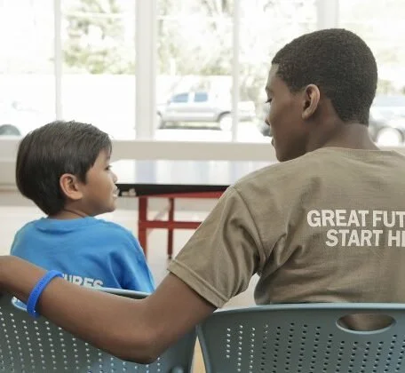 An older boy and younger boy sitting in a room, facing each other, with the older boy resting his arm on the back of the chair. The younger boy is wearing a blue shirt, and the older boy's shirt has the words 'GREAT FUTURE START HERE' printed on the back. There are large windows in the background letting in natural light.