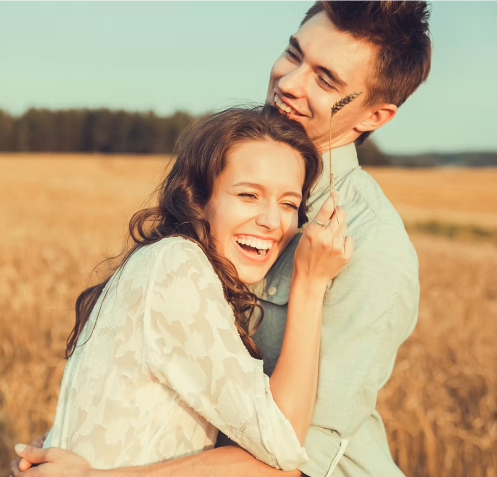 A smiling couple hugging outdoors in a field, with the woman holding a small plant in her hand.