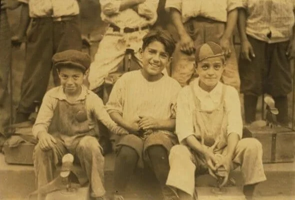 Three children are sitting in front of a group of people, smiling, in what appears to be an old black-and-white photo.