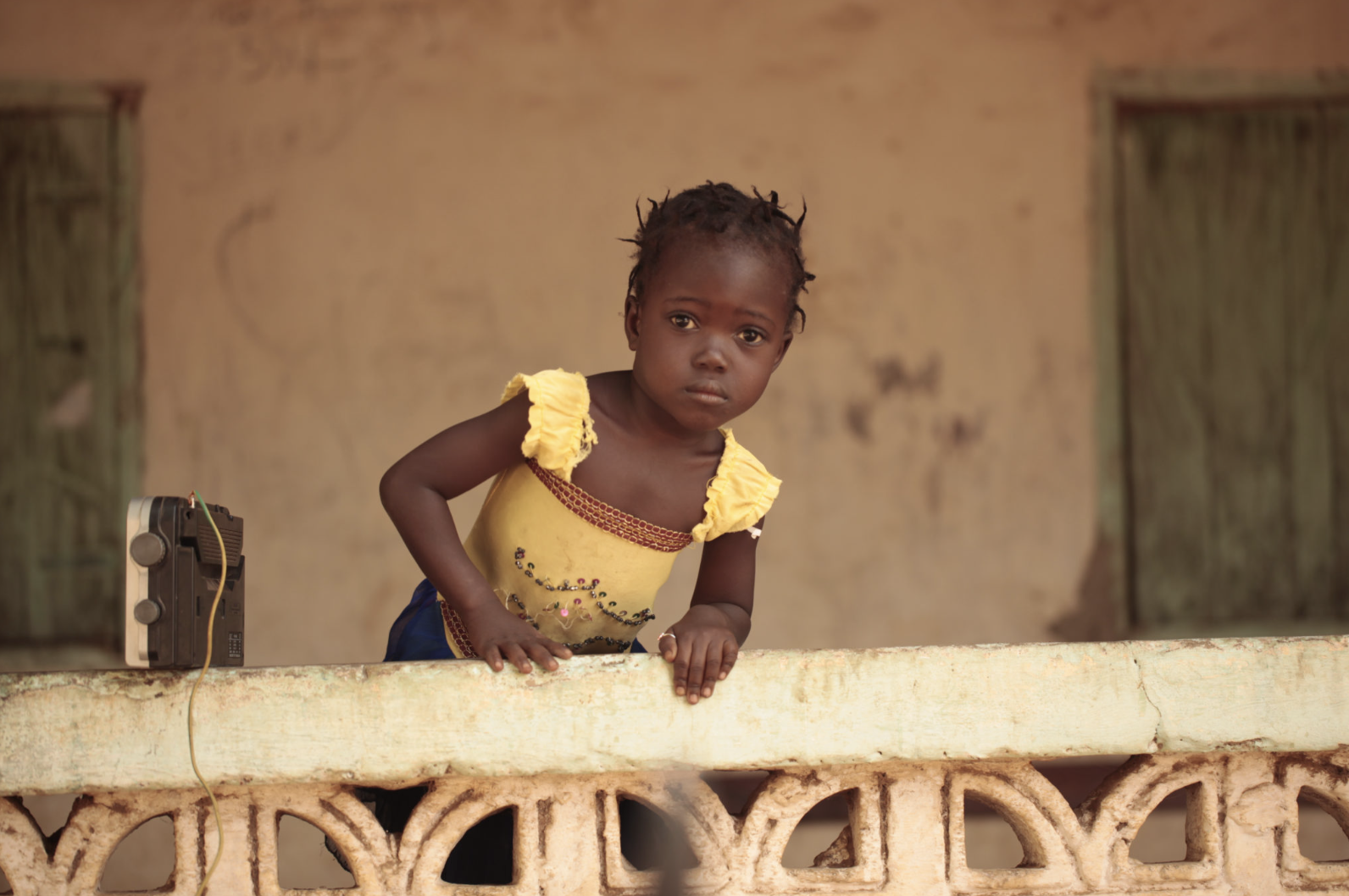 A young girl in a yellow top leaning over a concrete railing, with a background of a weathered wall and two green doorways.