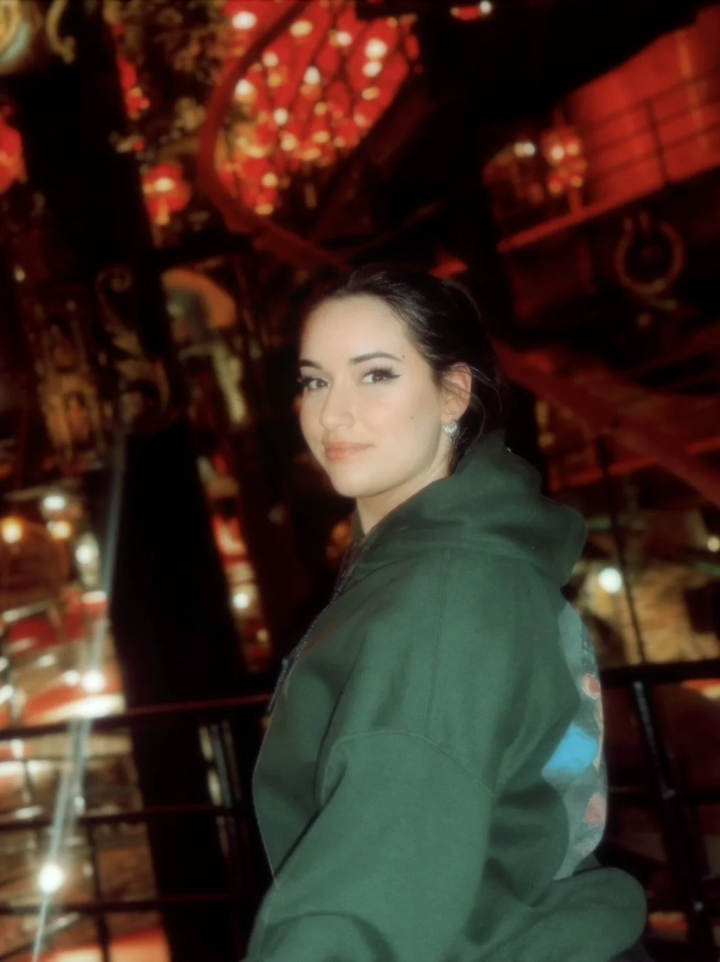 A woman with dark hair and earrings turns slightly and looks at the camera with a slight smile. She is wearing a green hoodie and is in a dimly lit interior space with red lights and decorative elements.