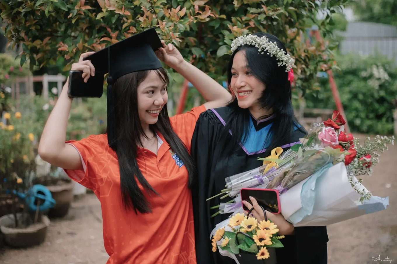 Two young women celebrating graduation outdoors. One is in a graduation cap and gown, holding a bouquet of flowers and a mobile phone. The other wears a floral headband and an orange shirt, adjusting the cap. They are smiling with a background of greenery and flowers.