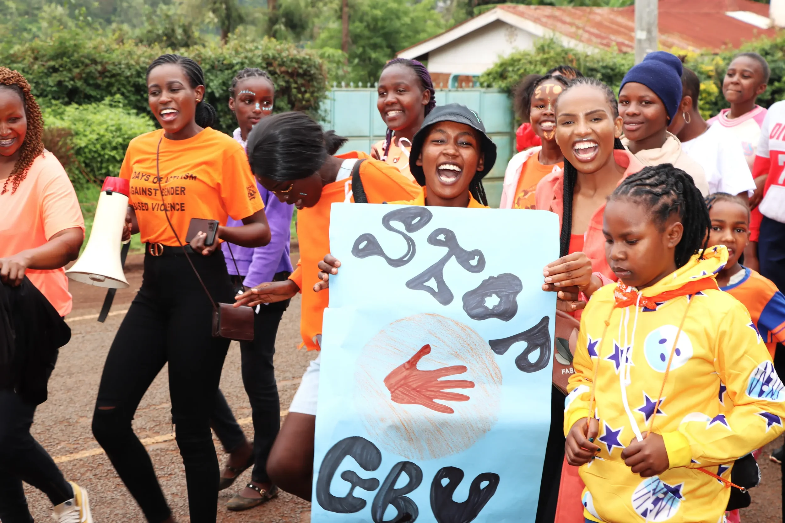A group of children and young adults participating in a community march or rally outdoors, some holding a sign and others engaged in conversation and smiling, with a background of trees, houses, and a fence.