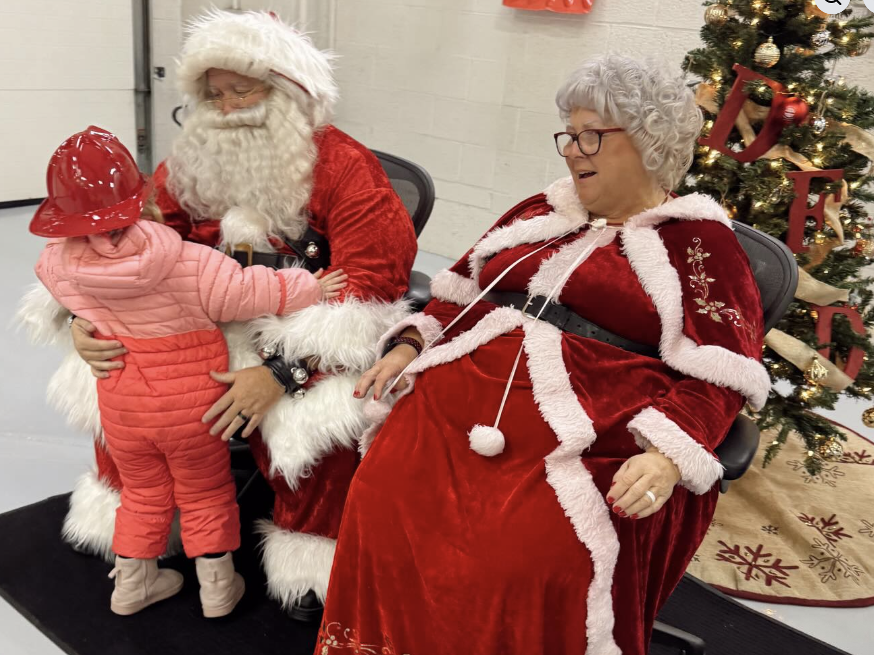A young girl hugging Santa Claus, who is sitting in a chair, while a woman dressed as Mrs. Claus sits nearby. There is a decorated Christmas tree with ornaments and lights in the background.