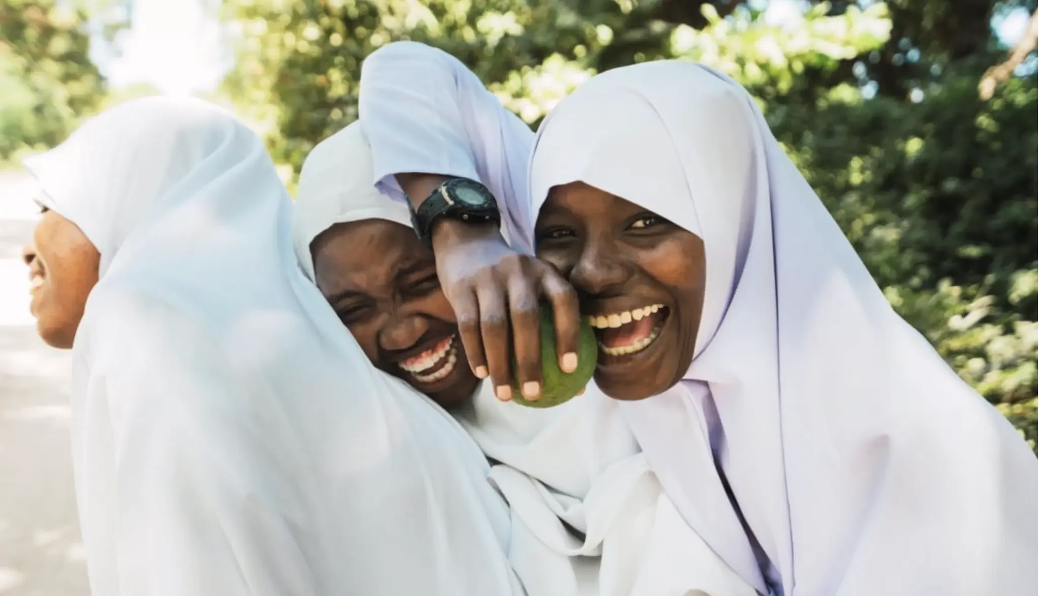 Two women wearing white headscarves are smiling and laughing while holding a green coconut. They are outdoors with blurred trees in the background.