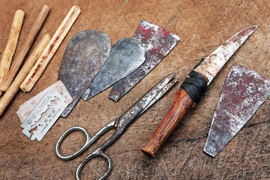 A collection of un-sanitized tools and knives on a wooden surface, including chisel blades, scissors, and a carving knife. Used for female genital mutilation.