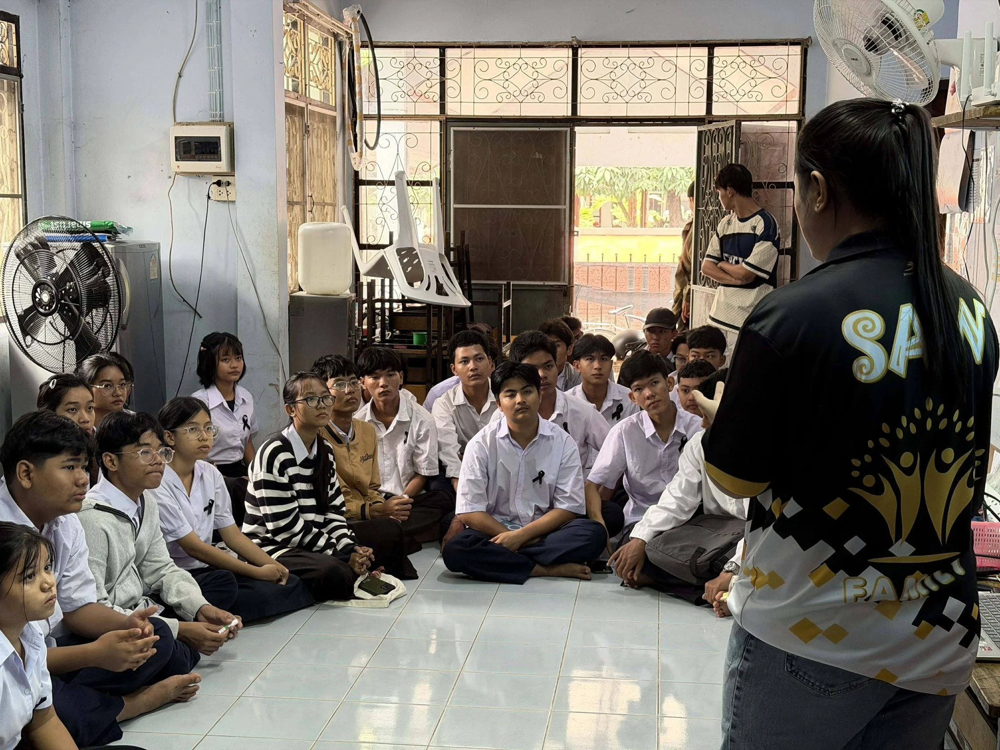 A group of students seated on the floor of a room, attentively listening to a woman standing in front of them. The woman is wearing a jacket with writing on the back and appears to be leading a discussion or giving a talk. There is a fan and a water dispenser in the room, with a window and metal grille in the background.