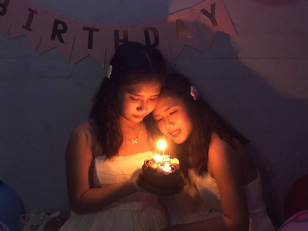 Two young women celebrate a birthday with a cake and candles, illuminated by candlelight in a dimly lit room decorated with a 'Happy Birthday' banner.