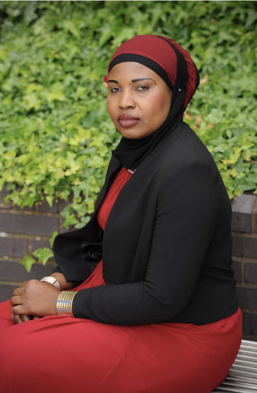 A woman survivor of FGM seated outdoors wearing a red dress, black blazer, and a hijab with red and black colors. She is sitting in front of a green leafy background with a brick wall. She has a neutral expression.