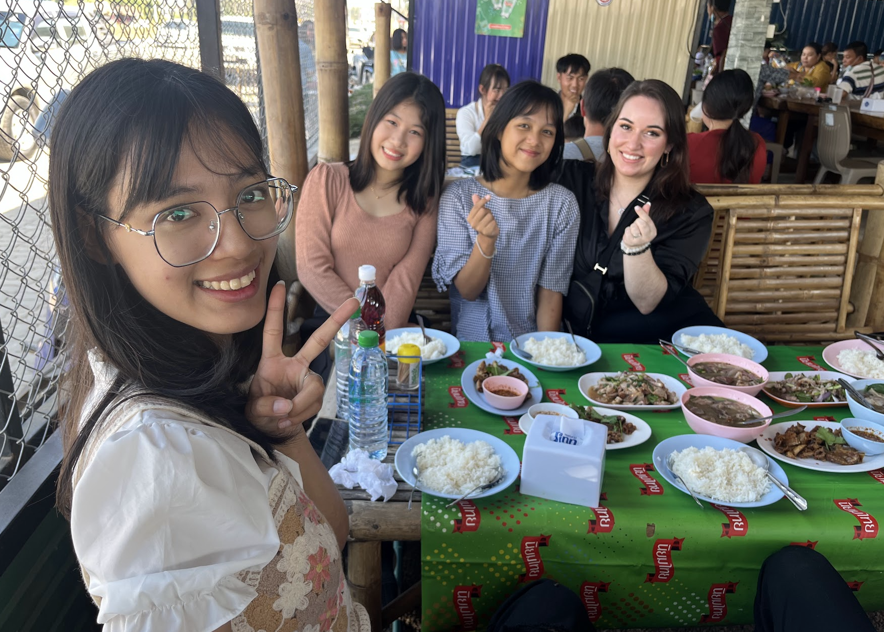 A group of five women sitting around a table at an outdoor restaurant, smiling and posing for the camera. The table is filled with bowls of rice, dishes of meat and vegetables, and drinks. Four women are in the background, and one woman with glasses and a peace sign is in the foreground.