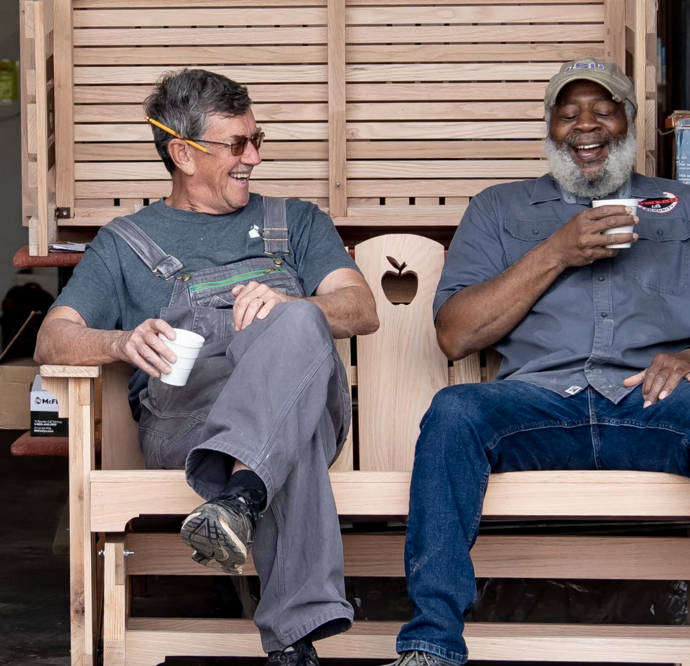 Two men sitting on a wooden bench, smiling and laughing, holding cups, with a wooden backdrop behind them.