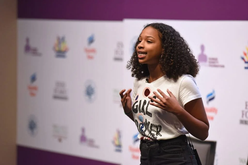 Young girl with curly hair speaking passionately at an event, standing in front of a backdrop with various logos.