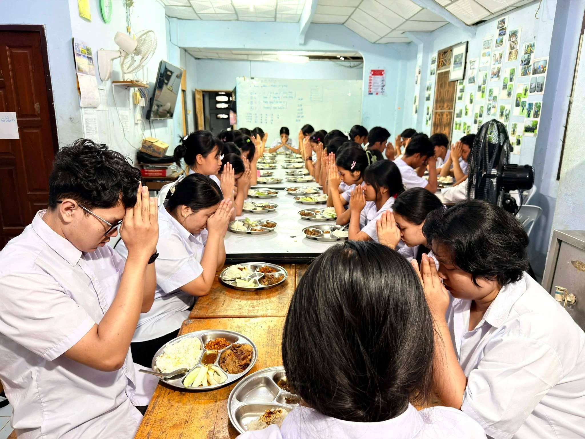 A group of students in school uniforms sitting at a long wooden table with plates of food, praying before eating in a classroom with light blue walls decorated with pictures and a whiteboard.